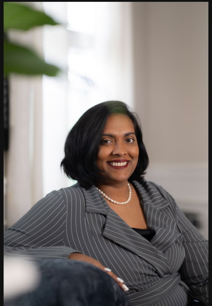 Portrait of Attorney Nisha Williams, smiling while seated in a professional setting, wearing a gray striped suit and pearl necklace.